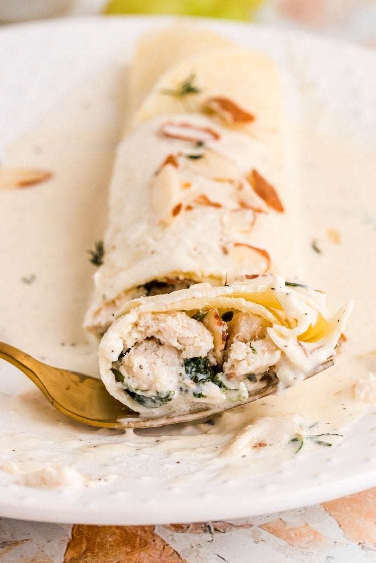 Close up photo of a spinach and chicken filled crepe with a gold fork taking a bite out of it from a white plate.