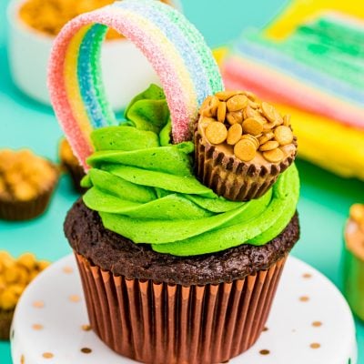 Chocolate cupcake with green frosting on a white cake stand. Decorated with rainbow and pot of gold candy..