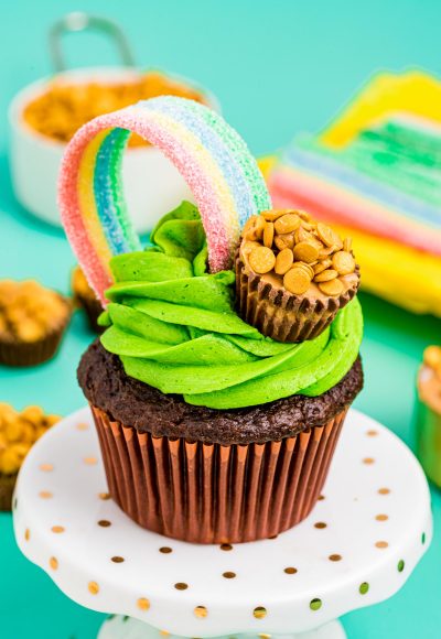 Chocolate cupcake with green frosting on a white cake stand. Decorated with rainbow and pot of gold candy..