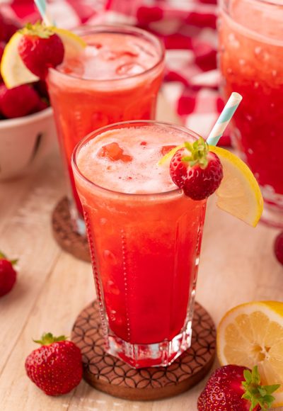 two glasses of strawberry lemonade on wooden coasters on a wooden table.