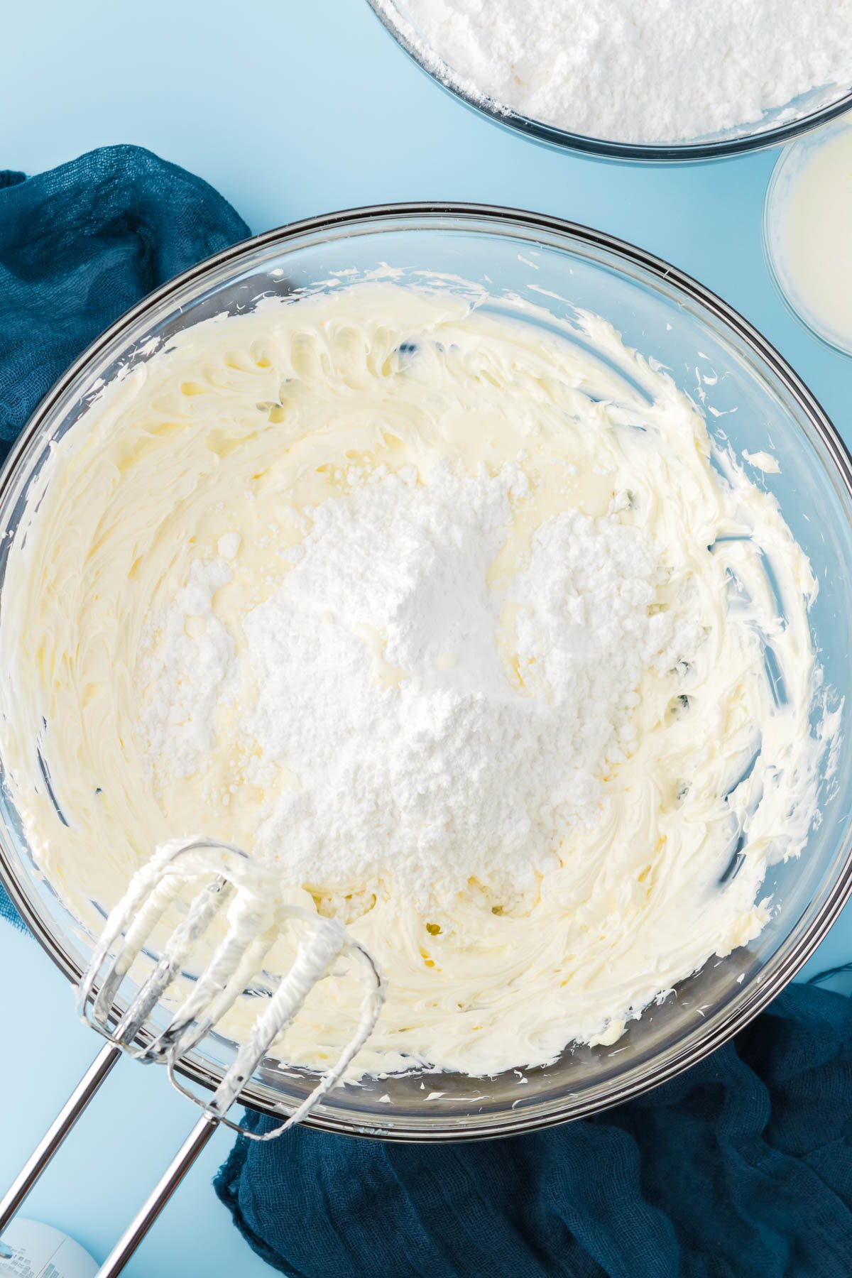 Powdered sugar being added to a glass bowl with creamed butter to make frosting.