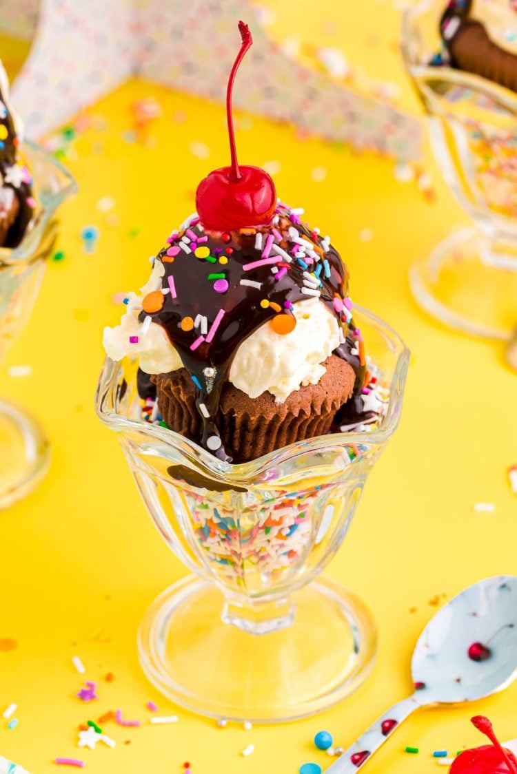 Close up photo of brownie cupcakes in a sundae dish filled with sprinkles on a yellow table.