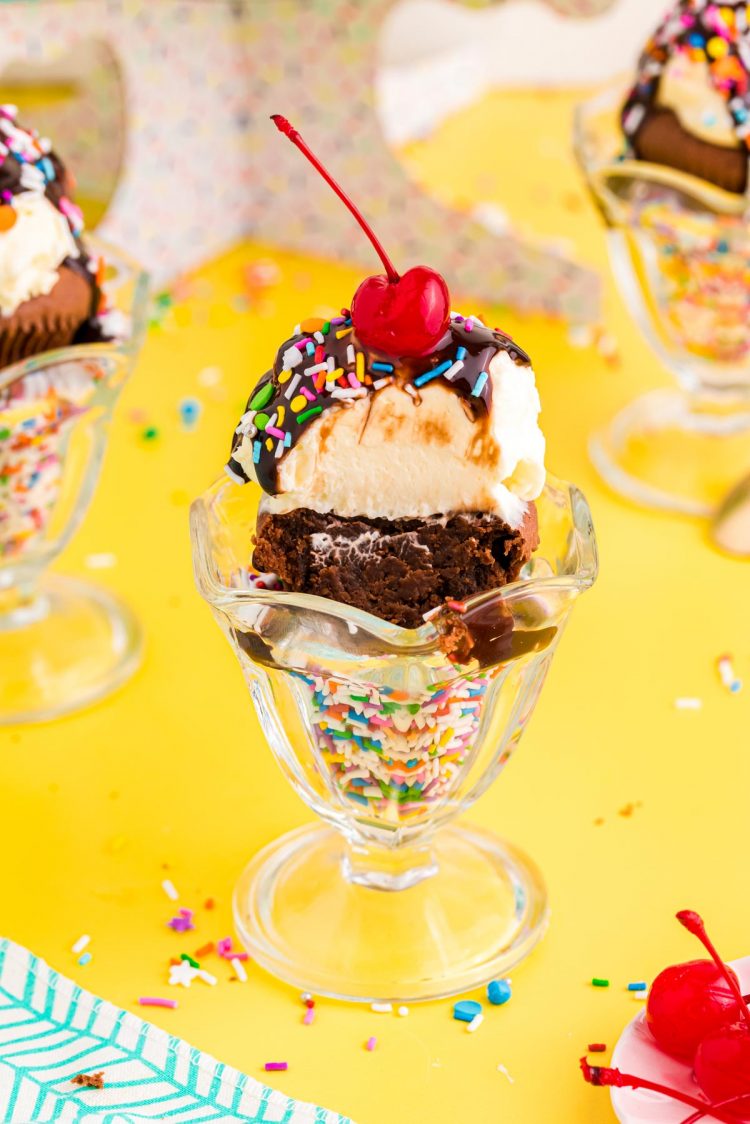 A brownie cupcake with a bite taken out of it in a sundae dish with rainbow sprinkles.
