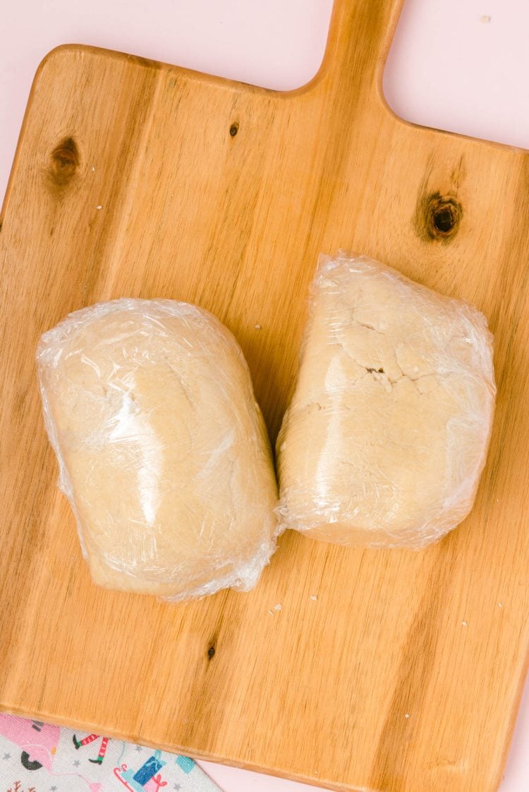Dough divided into two portions wrapped in plastic wrap on a wooden board.