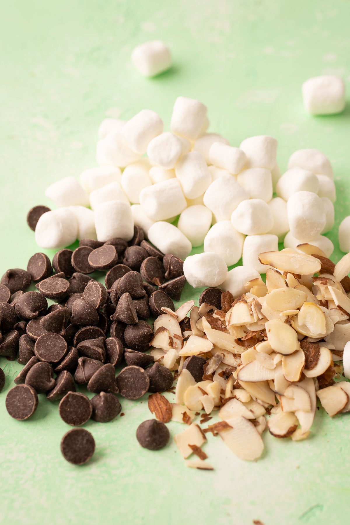 Chocolate chips, almond slivers, and mini marshmallows on a green table.