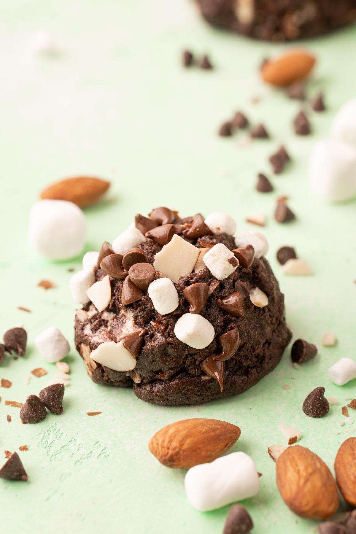 Rocky Road Cookie on a green surface with almond, marshmallows, and chocolate chips scattered around.
