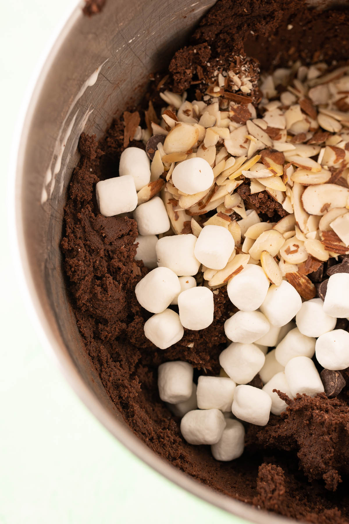 Chocolate cookie dough in a stainless steel mixing bowl with marshmallows, almonds, and chocolate chips being added in.