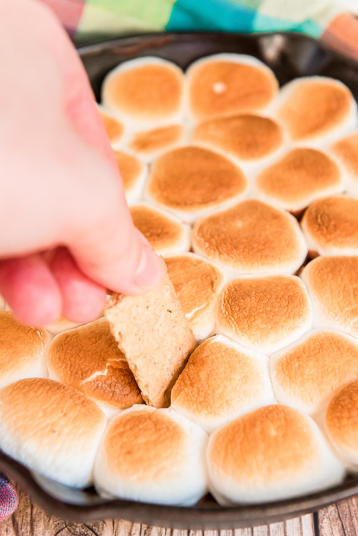 Woman's hand scooping a graham cracker piece into a cast iron skillet filled with toasted marshmallows.