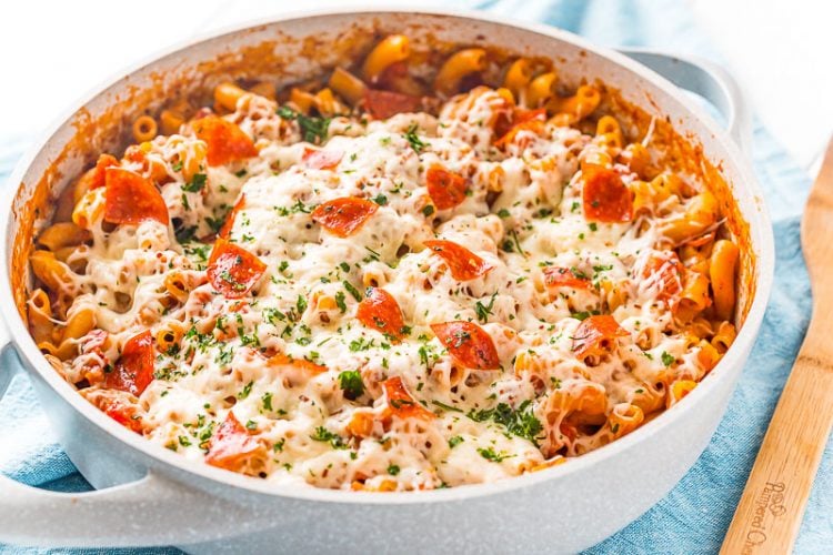 Pan filled with pizza pasta sitting on top of a blue napkin with a wooden spoon resting next to it.