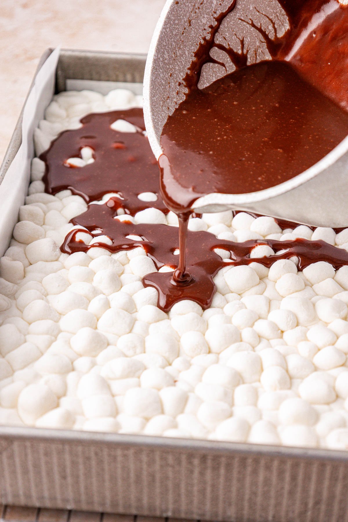 Hot chocolate icing being poured over marshmallows in a cake pan.