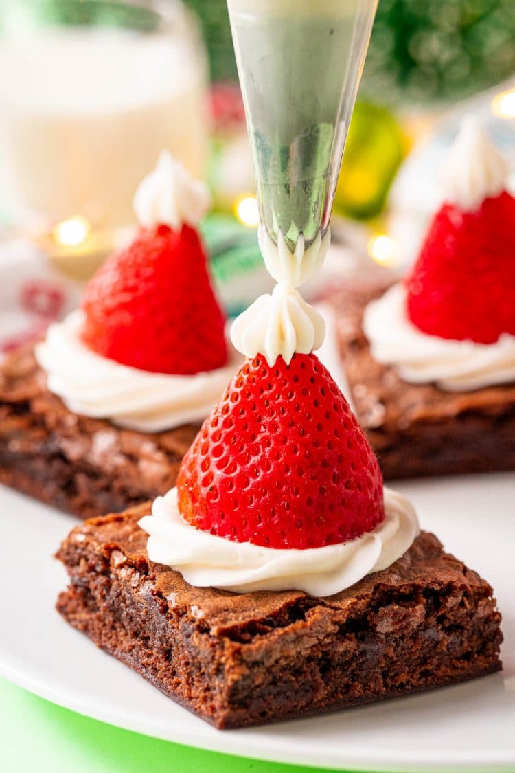 Buttercream frosting being piped onto the top of a strawberry on a brownie to make a santa hat.