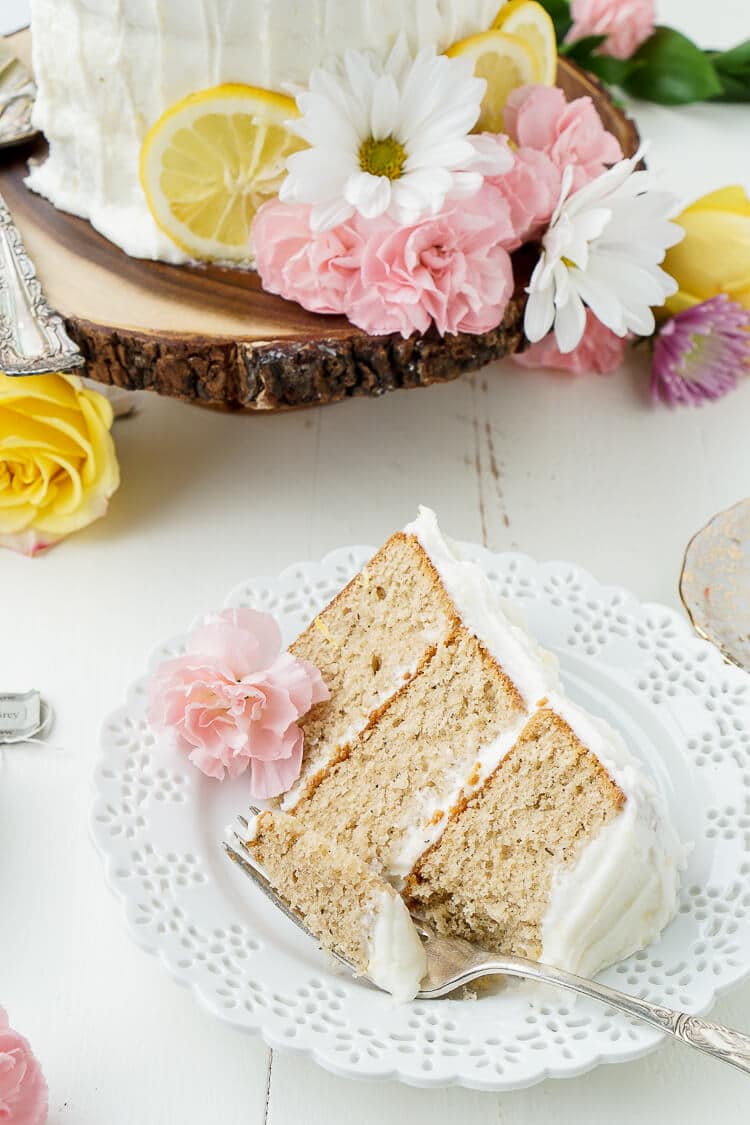 A close up of a slice of cake on a plate, with a cake, flowers and lemon slices in the background