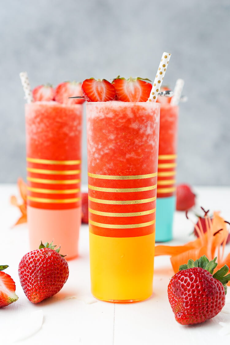 Strawberry Daiquiris in colorful glasses with fresh strawberries surrounding them on a white table with a grey backdrop.