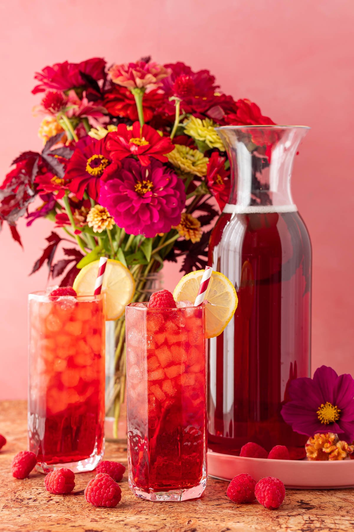 Glasses of raspberry iced tea on a table with a pitcher of tea and a vase of flowers.