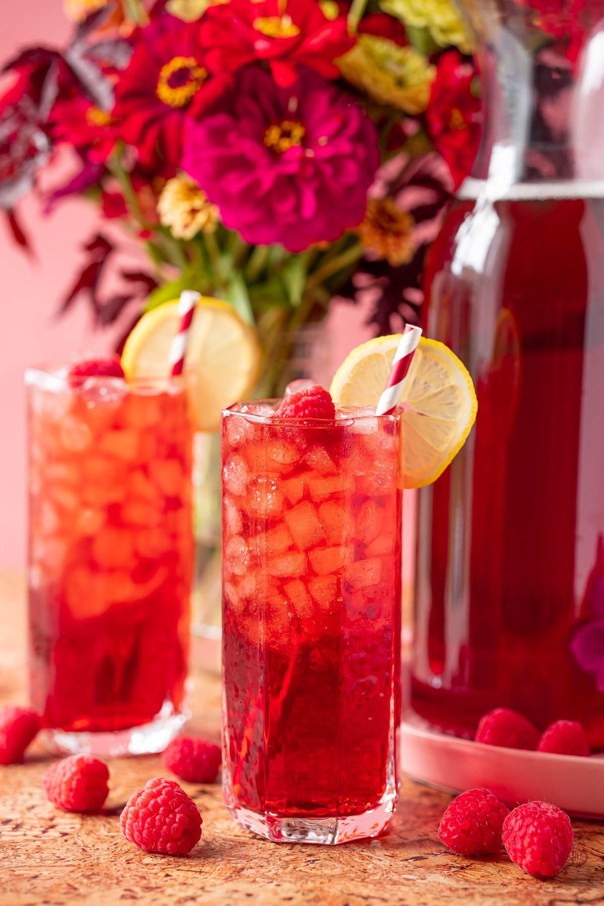 Two glasses filled with raspberry iced tea with flowers and a pitcher of tea in the background.