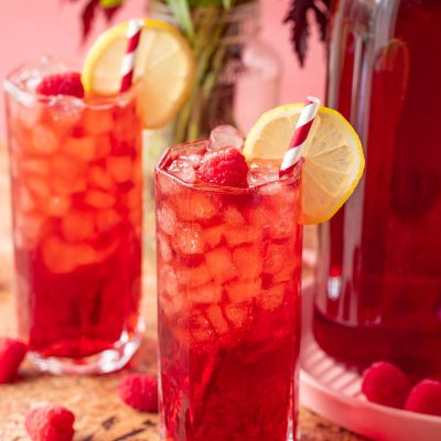 Two glasses filled with raspberry iced tea with flowers and a pitcher of tea in the background.