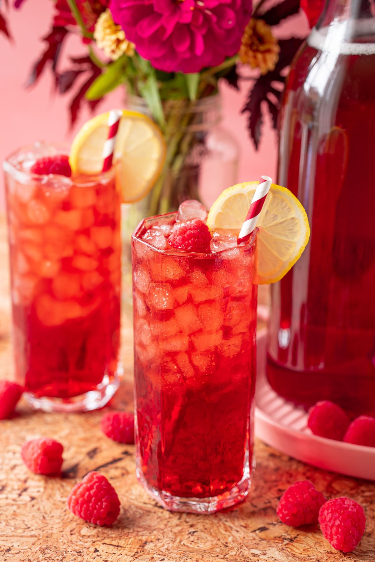 Two glasses filled with raspberry iced tea with flowers and a pitcher of tea in the background.