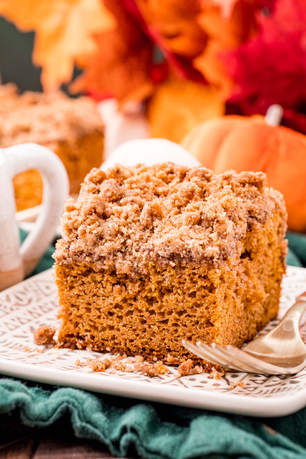 Close up photo of a slice of pumpkin coffee cake on a plate on a green napkin with fall decor in the background.