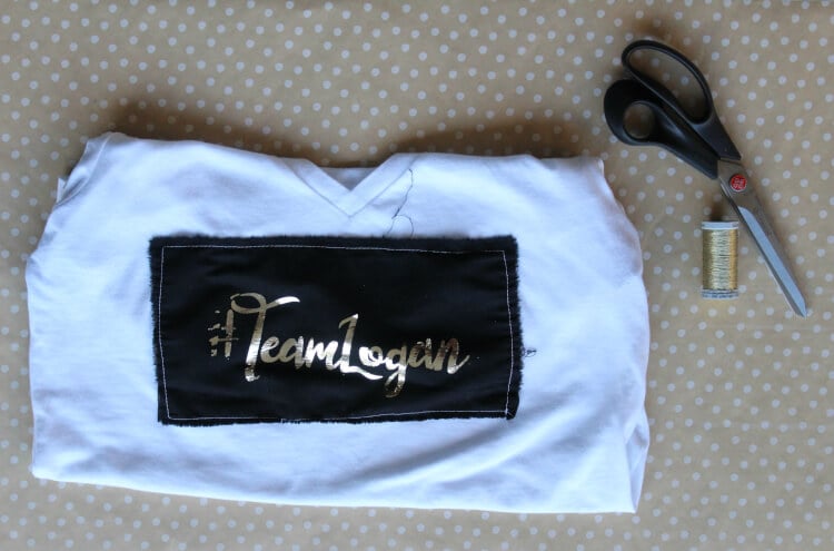 A scissors, a spool of thread and a blue shirt laying on a table and the shirt says, \"Team Logan\" 