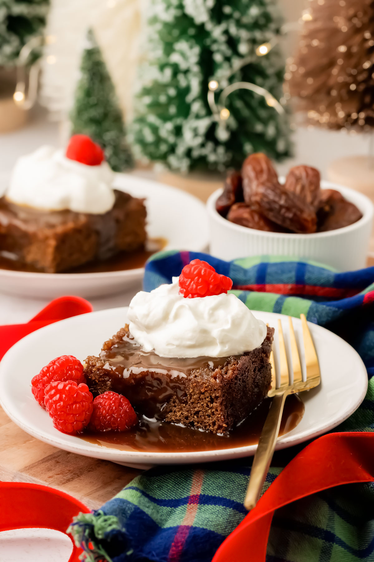 Slices of sticky toffee pudding on white plate with sauce, raspberries, and whipped cream.