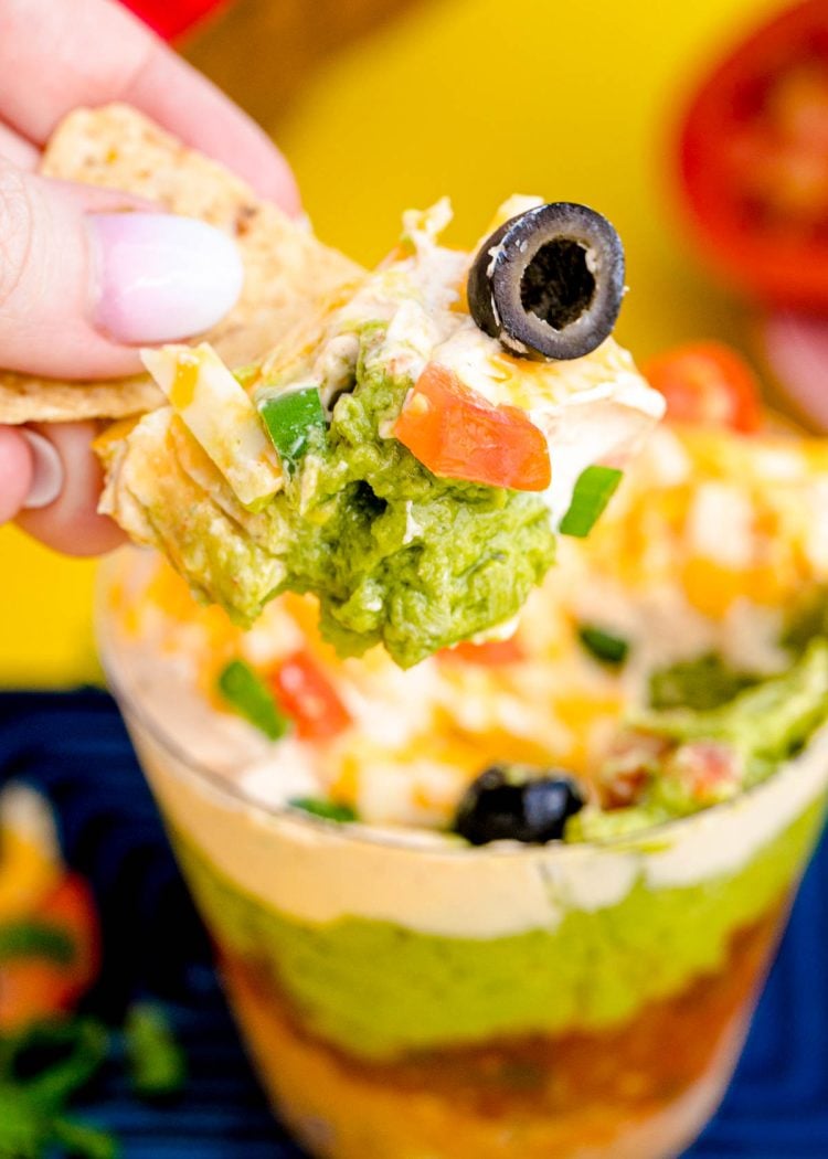 A woman's hand scooping a tortilla chip into a cup with 7 layer dip in it.