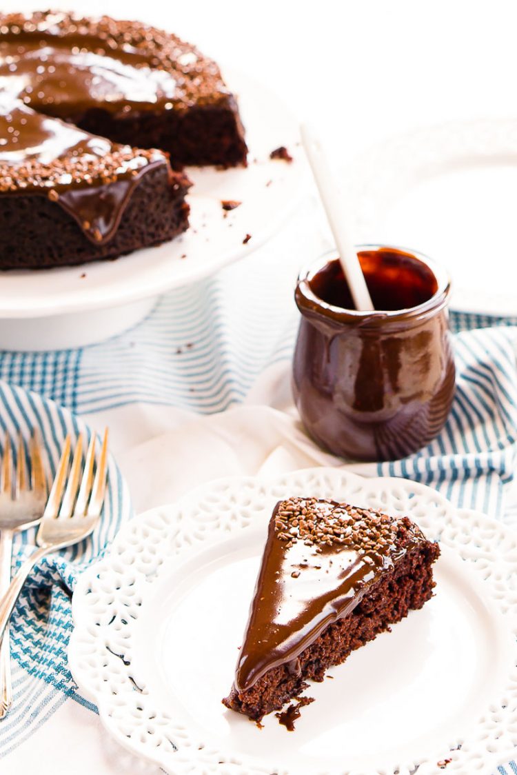 Slice of chocolate depression cake on a white plate with a jar of icing and the rest of the cake on a cake stand behind it.