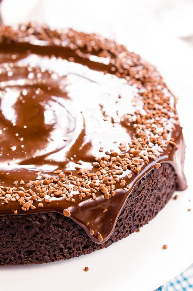 Chocolate Wacky Cake topped with chocolate icing and sprinkles on a white cake stand.