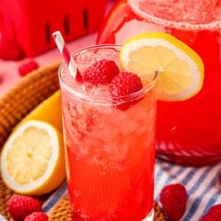 Close up of a glass and pitcher of raspberry lemonade on a blue and white striped towel.