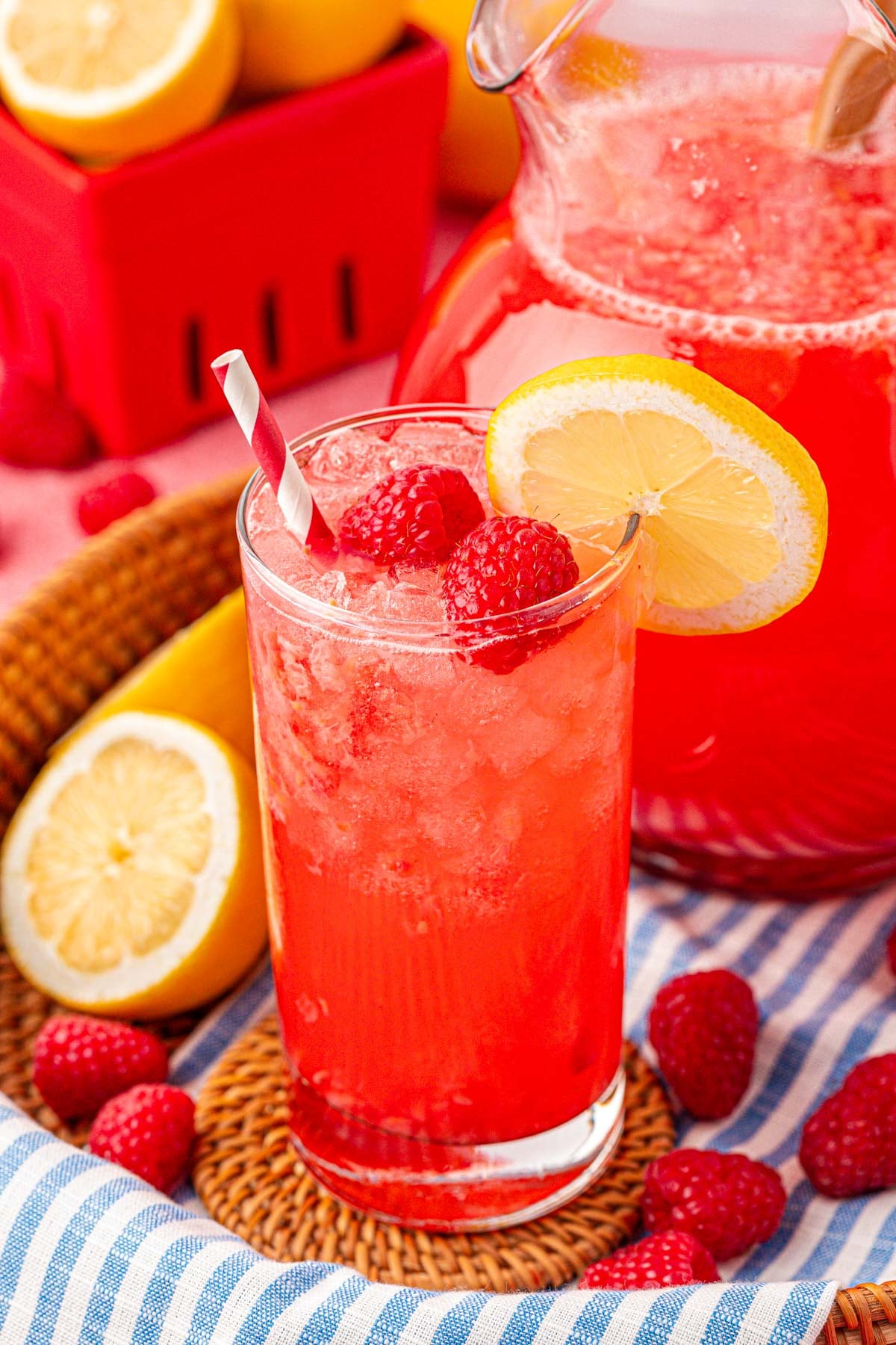 Close up of a glass and pitcher of raspberry lemonade on a blue and white striped towel.