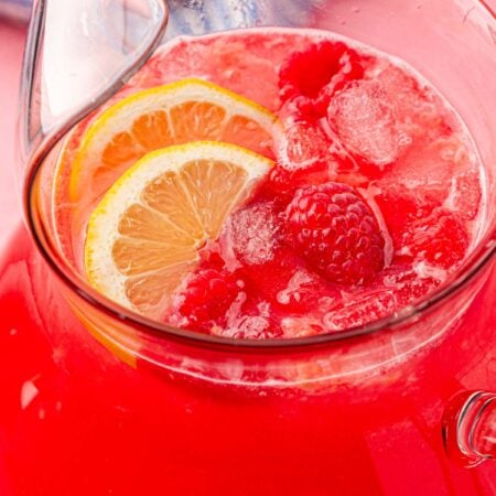 A pitcher of raspberry lemonade on a table with lemon slices and raspberries in it.