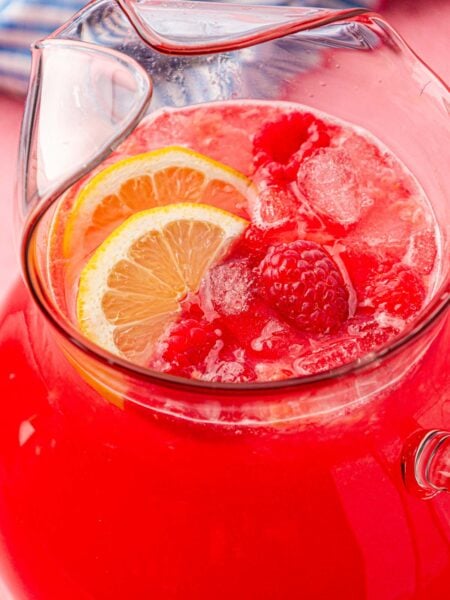 A pitcher of raspberry lemonade on a table with lemon slices and raspberries in it.