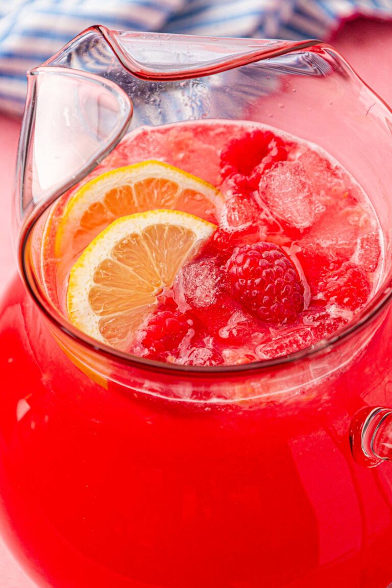 A pitcher of raspberry lemonade on a table with lemon slices and raspberries in it.