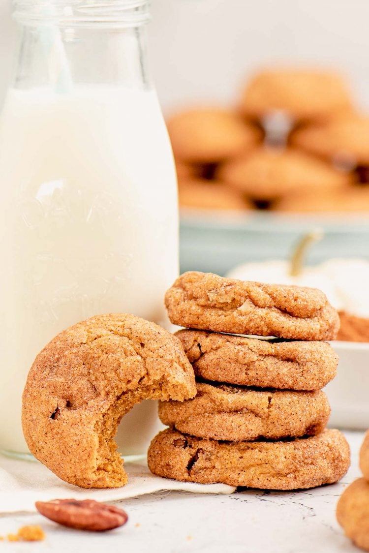 Close up photo of a stack of four pumpkin snickerdoodles next to a bottle of milk and one cookie with a bite taken out of it leaning against them.