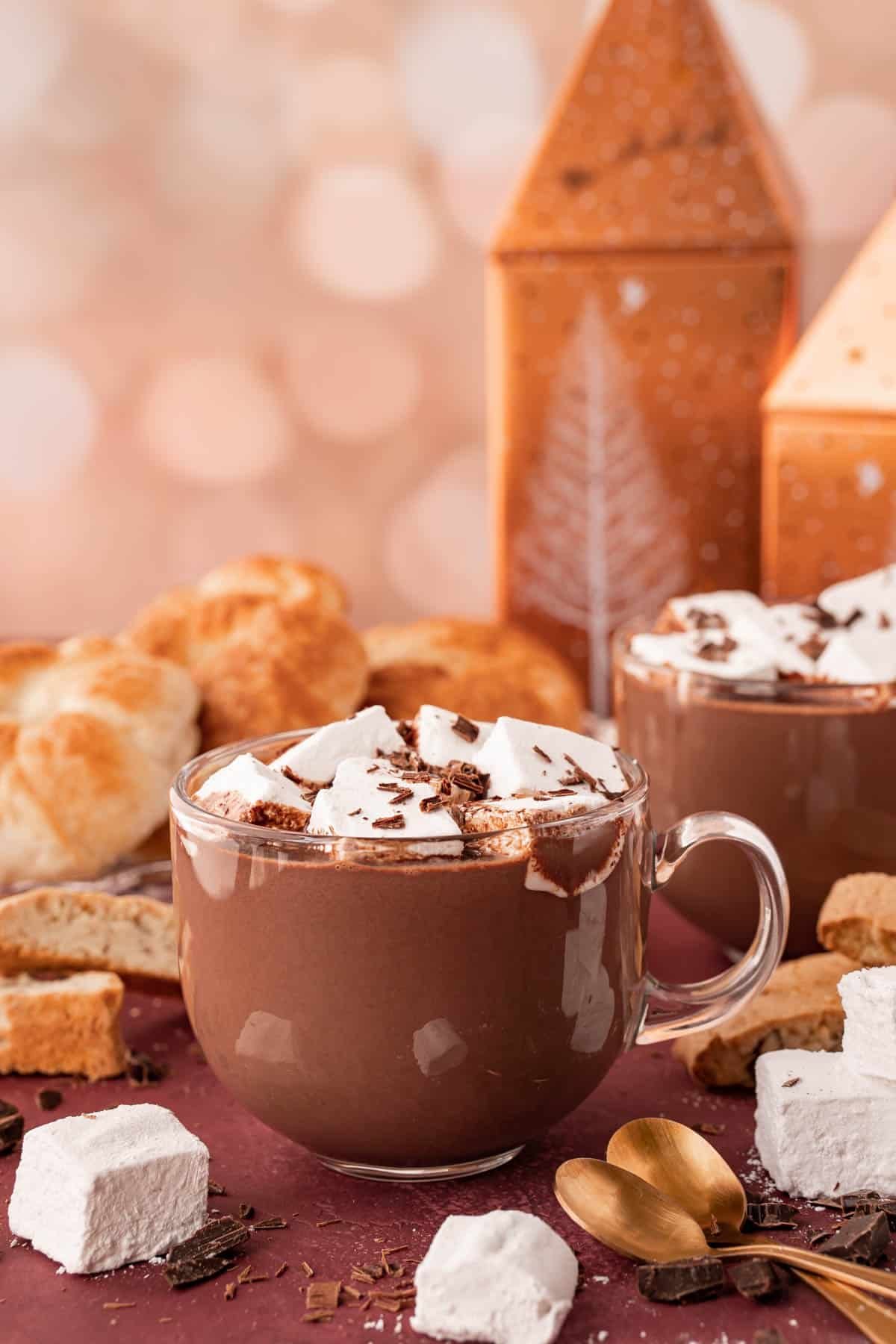 A clear glass mug filled with French Hot chocolate on a table with pastries and marshmallows around it.