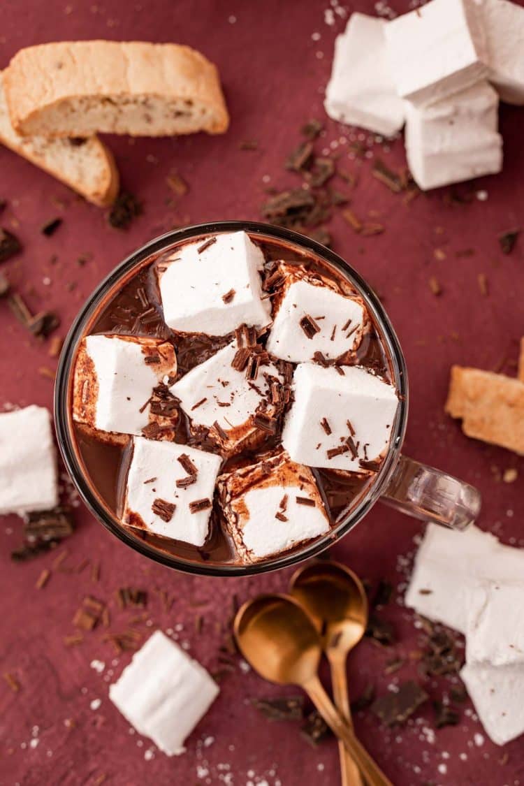 Overhead photo of a mug of French hot chocolate topped with marshmallows on a maroon surface.