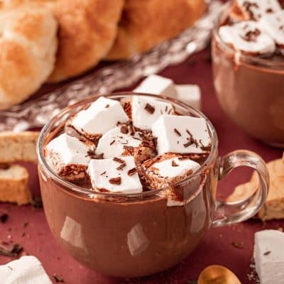 A clear glass mug filled with French Hot chocolate on a table with pastries and marshmallows around it.
