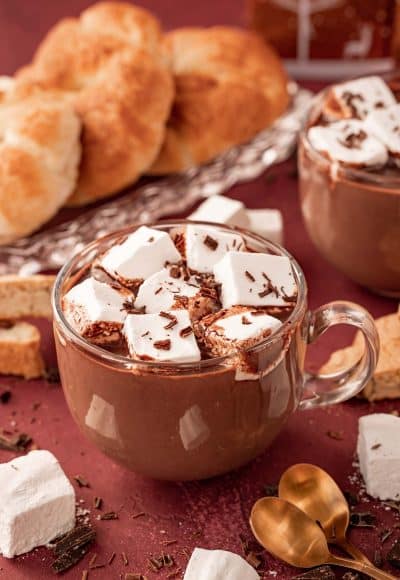 A clear glass mug filled with French Hot chocolate on a table with pastries and marshmallows around it.