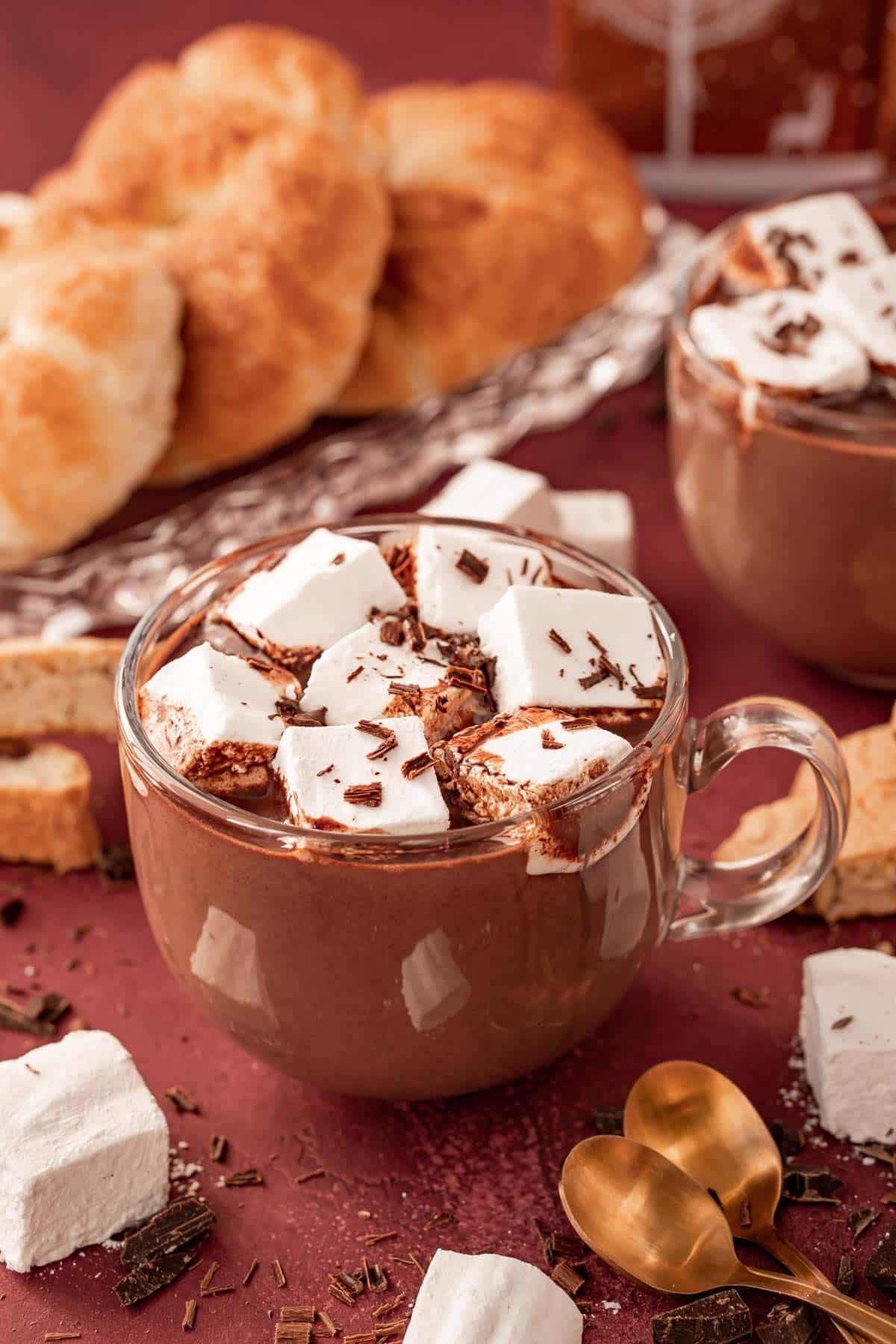 A clear glass mug filled with French Hot chocolate on a table with pastries and marshmallows around it.