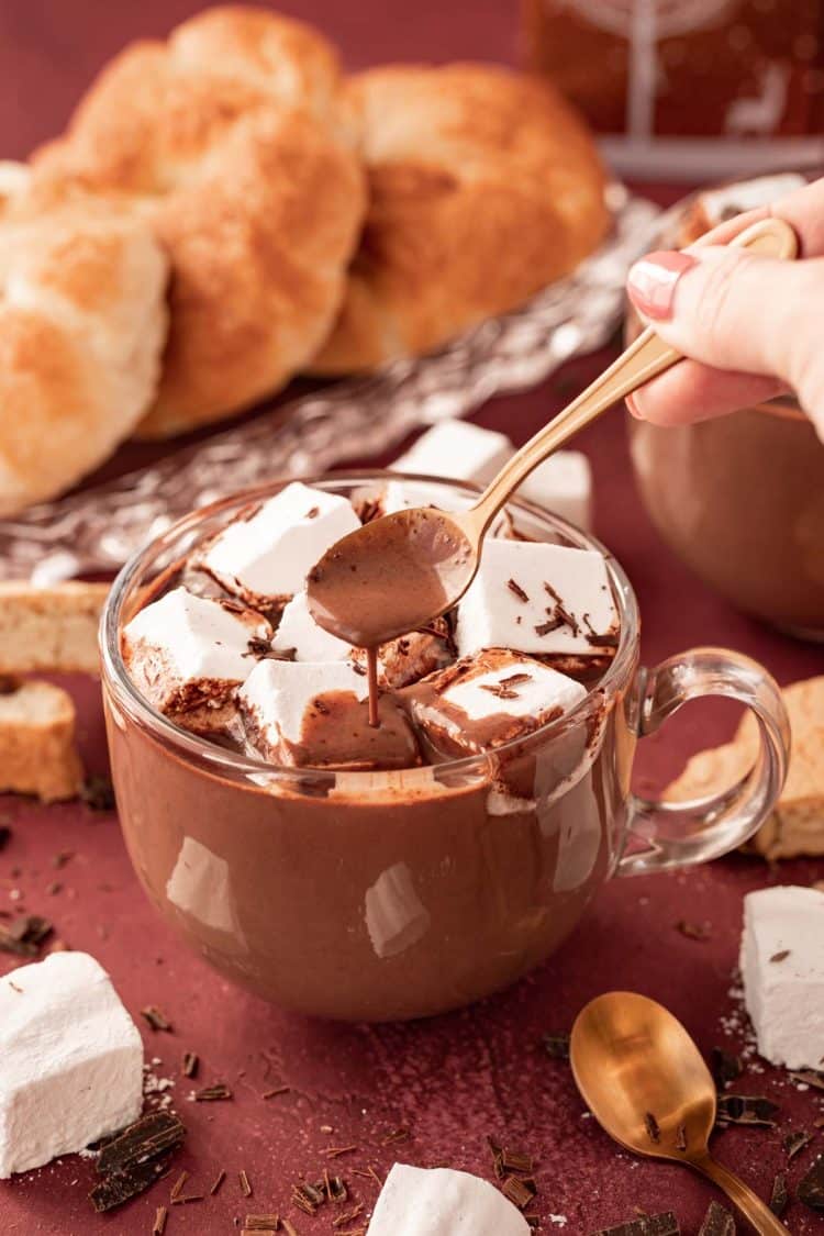 A clear glass mug filled with French Hot chocolate on a table with pastries and marshmallows around it. A spoon is dipping and drizzling the drink.