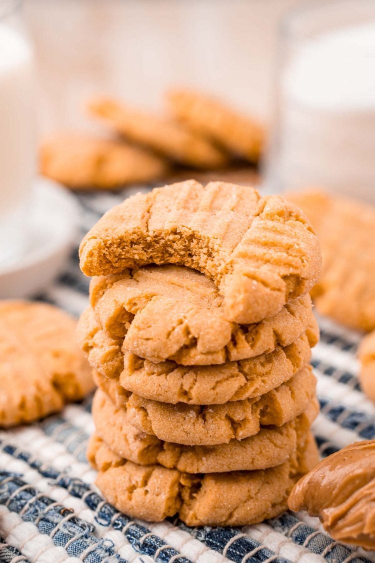 A stack of peanut butter cookies with the top cookie missing a bite on a striped napkin with more cookies and milk in the background.