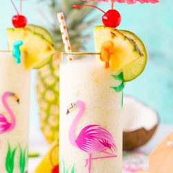 Glass with a flamingo painted on it with a pina colada in it topped with a drink umbrella and pineapple wedge.
