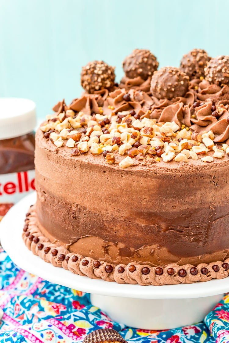 A chocolate cake made with nutella on a white cake stand topped with hazelnuts.
