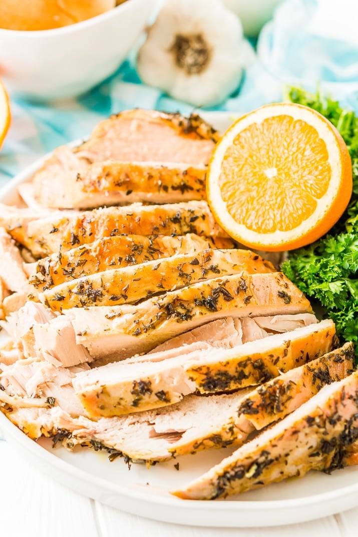 Sliced Turkey Breast with herbs on a white plate with half an orange, parsley, and garlic in the background.