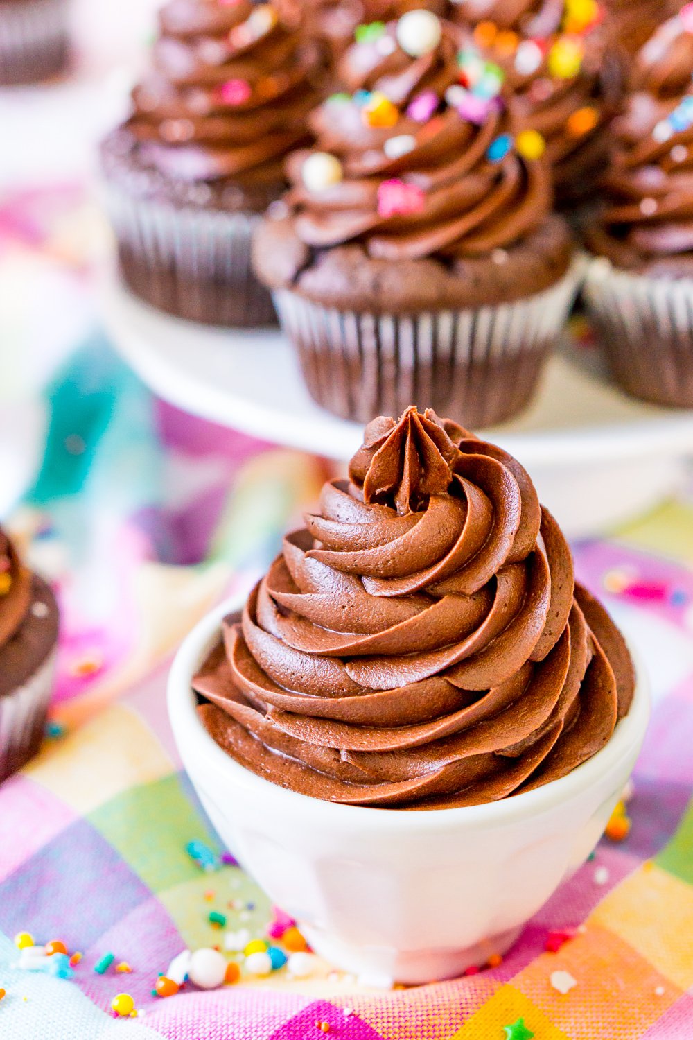 Chocolate Buttercream Frosting in white bowl with cake stand filled with chocolate cupcakes in background.