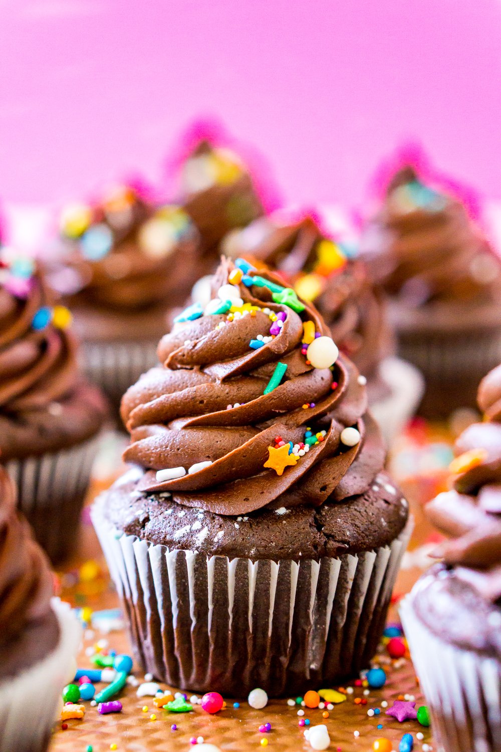 Chocolate Cupcakes on baking sheet with rainbow colored sprinkles and a pink background.