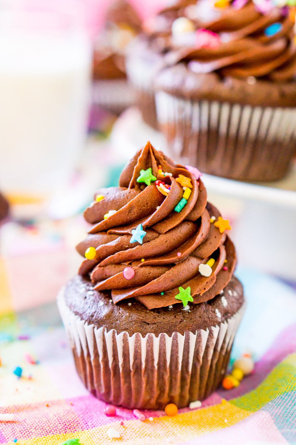 Chocolate Cupcake on colorful napkin with a white cake stand with more chocolate cupcakes in the background.