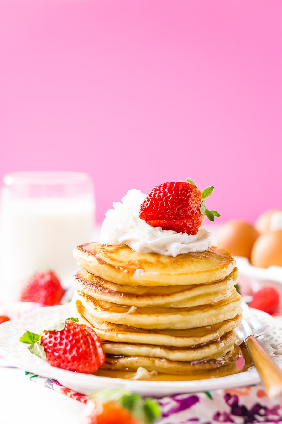Cream Cheese Pancake Stack on white plate with berries and whipped cream with pink background and floral napkin.