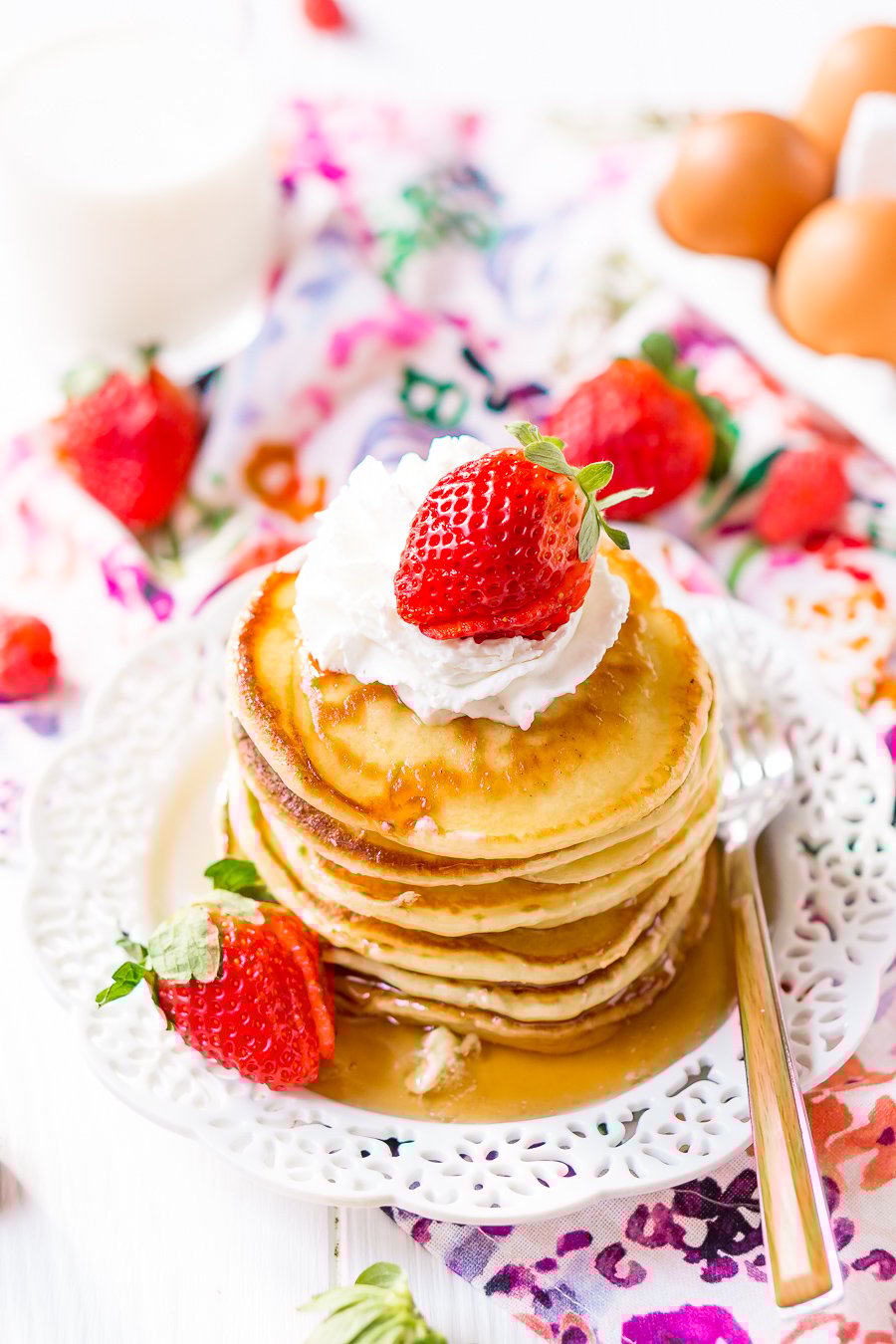 Stack of Cream Cheese Pancakes on whiter plate with whipped cream, maple syrup, and fresh strawberries.