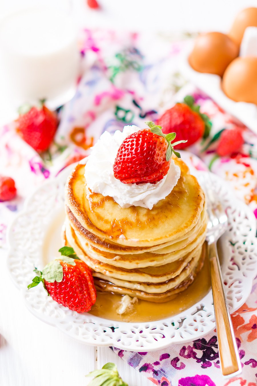 pilha de Cream Cheese Pancakes em prato mais branco com natas batidas, xarope de ácer e morangos frescos.