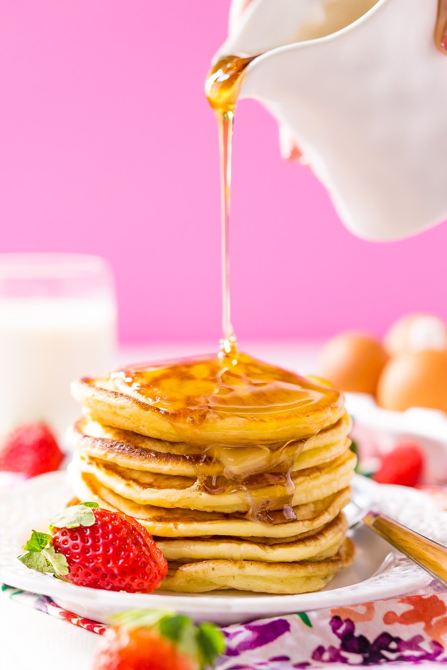 Pouring syrup over a stack of cream cheese pancakes on white plate with strawberries